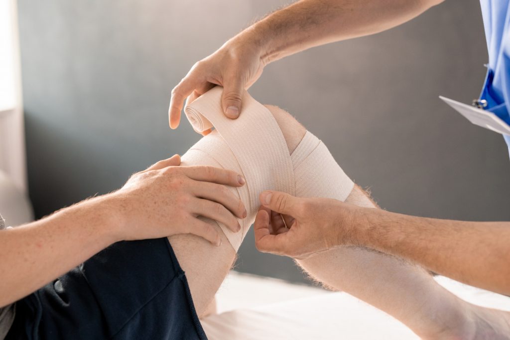 Hands of clinician wrapping right knee of patient with flexible bandage